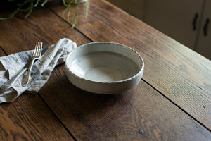 The coupe is a low bowl perfect for pasta, a salad, or anything saucy, with an elegant lip and a scalloped edge inspired by ceramics found in archaeological digs at the Historic 1747 Parsonage in Germantown, New York. Grey porcelain pasta bowl sitting on an old wood table with linen napkin.