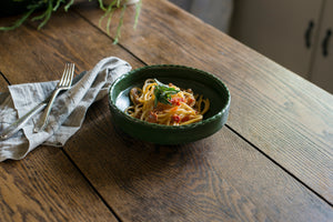 The coupe is a low bowl perfect for pasta, a salad, or anything saucy, with an elegant lip and a scalloped edge inspired by ceramics found in archaeological digs at the Historic 1747 Parsonage in Germantown, New York. Mugwort dark forest green porcelain bowl with bucatini pasta with tomato and basil. Linen napkin.