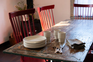 a wooden table with plates and cups on it. A porcelain dinner plate with an elegant lip and a scalloped edge, inspired by ceramics found in archaeological digs at the Historic 1747 Parsonage in Germantown, New York. Old house kitchen, silver tableware, linen napkin, old house dining room, windsor chairs, red chairs, old table, family dinner, dinner party