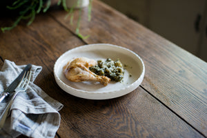 A porcelain dinner plate with an elegant lip and a scalloped edge, inspired by ceramics found in archaeological digs at the Historic 1747 Parsonage in Germantown, New York. Old house kitchen, silver tableware, linen napkin, chicken dinner, gnocchi, pesto, family dinner, dinner party