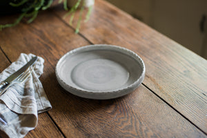 A porcelain dinner plate with an elegant lip and a scalloped edge, inspired by ceramics found in archaeological digs at the Historic 1747 Parsonage in Germantown, New York. Old house kitchen, silver tableware, linen napkin, grey porcelain plate, gray plate, smoke grey, porcelain plate, family dinner, dinner party