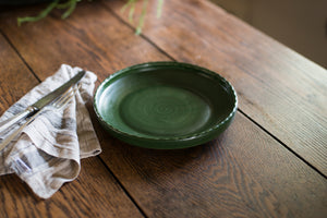A porcelain dinner plate with an elegant lip and a scalloped edge, inspired by ceramics found in archaeological digs at the Historic 1747 Parsonage in Germantown, New York. Old house kitchen, silver tableware, linen napkin, mugwort green, dark green, forest green, family dinner, dinner party