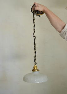 Hand holding a pendant light with a white glass shade and brass chain against a plain background