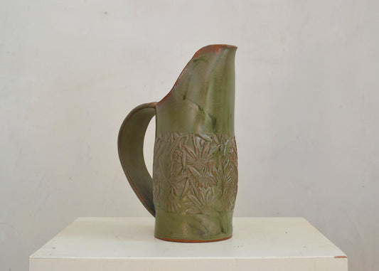 Green ceramic pitcher with decorative patterns on a white pedestal against a plain background