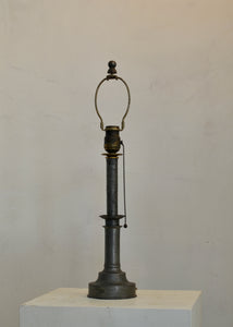 Antique lamp with metal base and brass fitting on a white pedestal against a plain background. Quittner