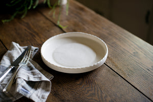 A porcelain dinner plate with an elegant lip and a scalloped edge, inspired by ceramics found in archaeological digs at the Historic 1747 Parsonage in Germantown, New York. Old house kitchen, silver tableware, linen napkin, white porcelain plate, family dinner, dinner party